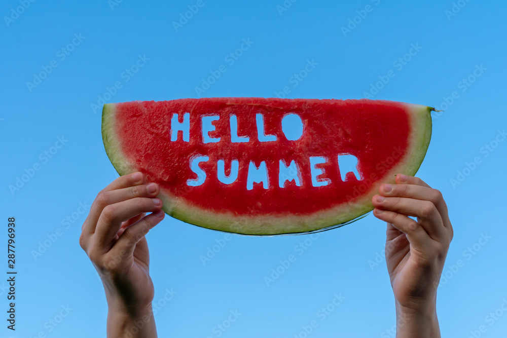 A piece of watermelon against a blue sky. Children's hands are holding a slice of watermelon with the text Hello Summer. Summer time concept - obrazy, fototapety, plakaty 