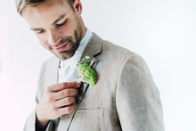 Handsome Bridegroom In Suit Holding Floral Boutonniere And Looking At It Isolated On Grey