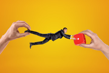 Poster - Big male hand holding tiny businessman who is reaching to another big hand holding red casino dice on yellow background