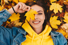Autumn Walk. Woman Portrait. Happy Girl In Yellow Hoodie And Jean Jacket Is Playing With Leaf, Looking At Camera And Smiling While Lying On The Ground In The Park; Top View