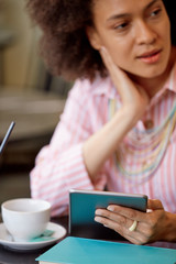 Wall Mural - Attractive mixed race woman in striped pink dress sitting in cafe and using tablet.