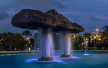 In The Evening In The Public Park Of Nations In The Spanish City Of Torrevieja. A Fountain Has Blue Water Fountains And The Park With Large Expanses Of Water Is Brightly Lit.