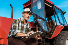 Senior Farmer In Straw Hat Using Digital Tablet Near Red Tractor