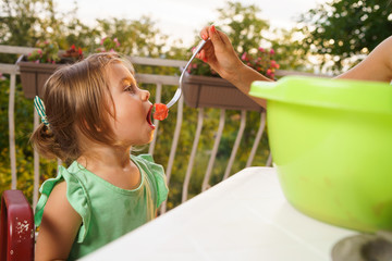 Portrait of little small girl blonde hair eating fruits watermelon in summer day by the table on her balcony fed by her mother