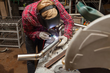 Wall Mural - Young guy welder in a checkered red shirt welds a stainless steel pipe using agronomic welding to protect his eyes with a mask in an iron workshop. Modern welding methods.