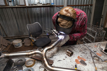 Canvas Print - Young guy welder in a checkered red shirt welds a stainless steel pipe for car exhaust using agronomic welding to protect his eyes with a mask in an iron workshop. Modern welding methods.