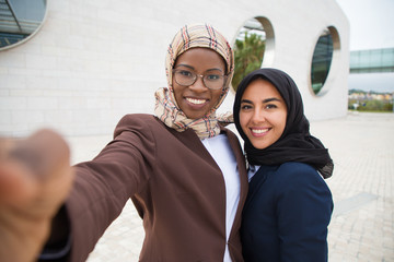 Wall Mural - Joyful female corporate friends taking selfie outside. Muslim business women in hijabs holding mobile phone, hugging each other, posing and smiling at screen. Business and friendship concept