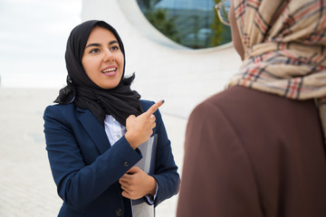 Wall Mural - Excited Muslim female employee consulting colleague outside, pointing finger away and speaking. Business women in hijabs standing outdoors and discussing project. Muslim female workers concept