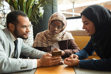 Wall Mural - Excited diverse coworkers watching content on smartphone. Businessman and Muslim businesswomen sitting in cafe, man showing phone screen to colleagues. Digital technology concept