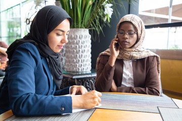 Wall Mural - Serious female business colleagues working on project in coffee shop. Muslim business women in hijabs sitting in cafe, talking on mobile phone and taking notes. Working in cafe concept