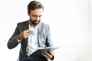 Wall Mural - Handsome optimistic smiling bearded business man in office drinking coffee holding clipboard.