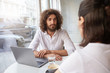 © timtimphoto - Indoor shot of handsome curly guy with beard sitting in cafe with business partner, having laptop and glass of water on table, looking attentively on person next to him