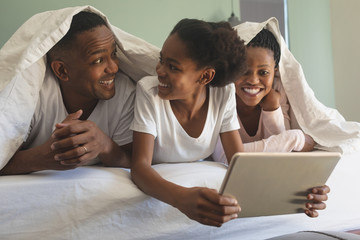 Wall Mural - Happy African American family under blanket and using digital tablet on bed in bedroom