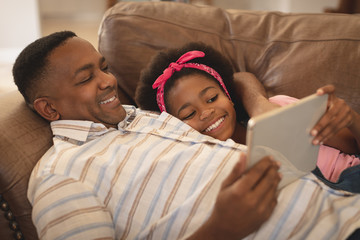 Wall Mural - Happy African American father and daughter relaxing on the sofa and using digital tabl