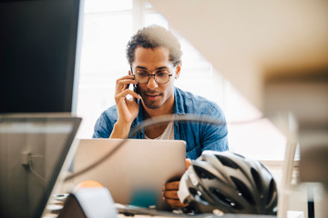 Computer programmer talking on smart phone while using laptop in creative office