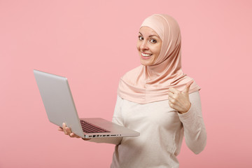 Smiling young arabian muslim woman in hijab light clothes posing isolated on pink background. People religious lifestyle concept. Mock up copy space. Hold and work laptop pc computer showing thumb up.