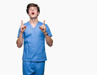 Wall Mural - Young doctor wearing medical uniform over isolated background amazed and surprised looking up and pointing with fingers and raised arms.