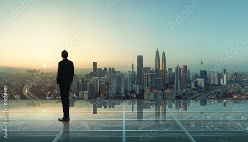 Fotomural Businessman standing at transparent glass floor on rooftop with city skyline, success and thinking concept