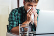 © GENETTICA - Glass of water and pill on the office table to relieve symptoms of cold, flu or allergy. Young man sitting in front of the computer blowing his nose in the background.