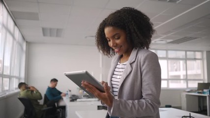 Poster - Successful smiling young african businesswoman in grey jacket using a digital tablet in front of colleague working at workplace