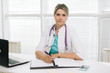 © dikushin - Portrait of young woman doctor in hospital office with laptop taking patient. Female therapist sits at the table in front of the camera and looking at camera