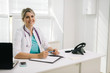 © dikushin - Portrait of young smiling woman doctor in hospital office with laptop taking patient. Female therapist sits at the table in front of the camera and looking at camera