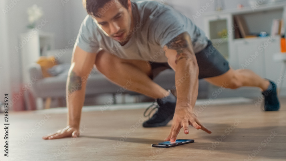 Muscular Athletic Fit Man in T-shirt and Shorts is Doing Mountain Climber Exercises While Using a Stopwatch on His Phone. He is Training at Home in His Bright Living Room with Minimalistic Interior. - obrazy, fototapety, plakaty 
