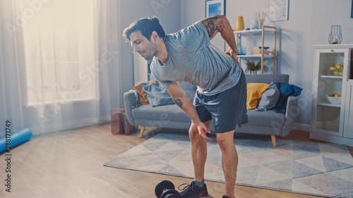 Cuadros en Lienzo Muscular Athletic Shirtless Fit Man in Grey Shorts Injures His Back After Lifting Dumbbells at Home in His Spacious and Sunny Living Room with Minimalistic Interior