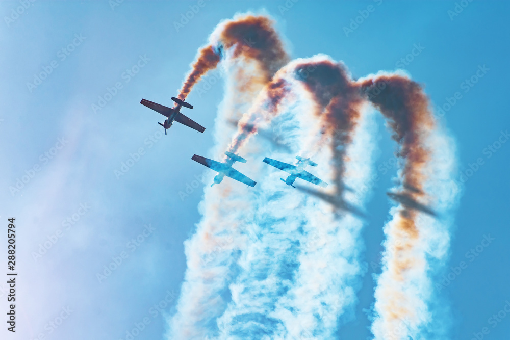 Three light-engine aircraft perform aerobatics - a dead loop. The bright sun illuminates the planes and the shadows fall on the smoke they leave in the sky. - obrazy, fototapety, plakaty 