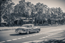Black And White Monochrome Photo Of An American Oldtimer Car From The 1950s. Vintage USA Car On The Street On A Sunny Summer Day. Retro Travel, Traffic Concept.
