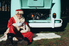 Senior Man In Costume Of Santa Claus Speaking On Mobile Phone While Sitting By Van With Open Engine Compartment