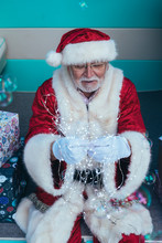 Senior Man In Costume Of Santa Claus Sitting In Retro Van Between Presents And Holding Garland In Gloved Hands