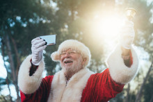 From Below Joyful Man In Costume Of Santa Claus Ringing Bell And Taking Selfie With Mobile Phone On Blurred Nature Background