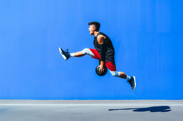 Side view of a young man holding a basket ball jumping in front of a blue wall.