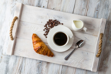 Wall Mural - A white Cup of coffee with a croissant on a light wooden tray top view