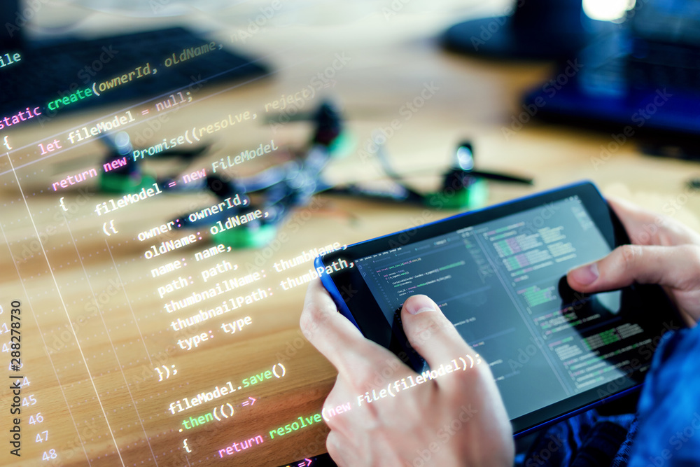 Programmer is holding tablet with program code of software for controlling FPV drone. Developer is building quadcopter from kit with microcontrollers at workplace. Desktop of hardware engineer.