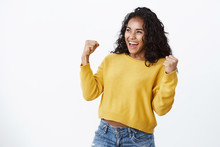 Lucky Venturous Young African-american Girl Fan In Yellow Sweater, Making Fist Pump Gesture As Smiling And Looking Left, Celebrating Team Scored Goal, Achieve Success, Winning Huge Money