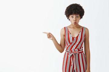 Wall Mural - Portrait of shocked and impressed girlfriend asking question about guy got rish suddenly, pointing left and folding lips from curiousity and amazement being under great influence over gray wall