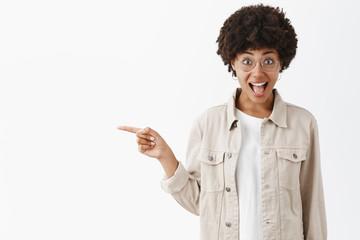 Wall Mural - Portrait of happy emotive and expressive young dark-skinned woman in beige trendy shirt and glasses pointing left with forefinger and smiling joyfully, entertained and amused over gray background