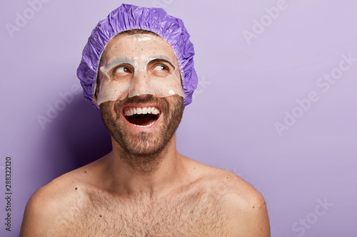 Fotografía Happy positive young man applies facial mask, wears shower cap, laughs positively, has naked body, dark bristle, enjoys spa treatment, isolated over purple background, feels relaxed