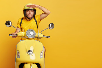 Concentrated male driver in helmet on scooter, keeps palm near forehead, poses on motorbike, carries rucksack, has serious face expression, poses against yellow background, copy space on right side