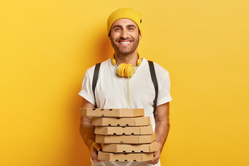 Wall Mural - Joyful cheerful man in casual wear, holds pile of cardboard boxes with pizza, has friendly expression, uses headphones for listening audio track, delivers junk food, demonstrates good service