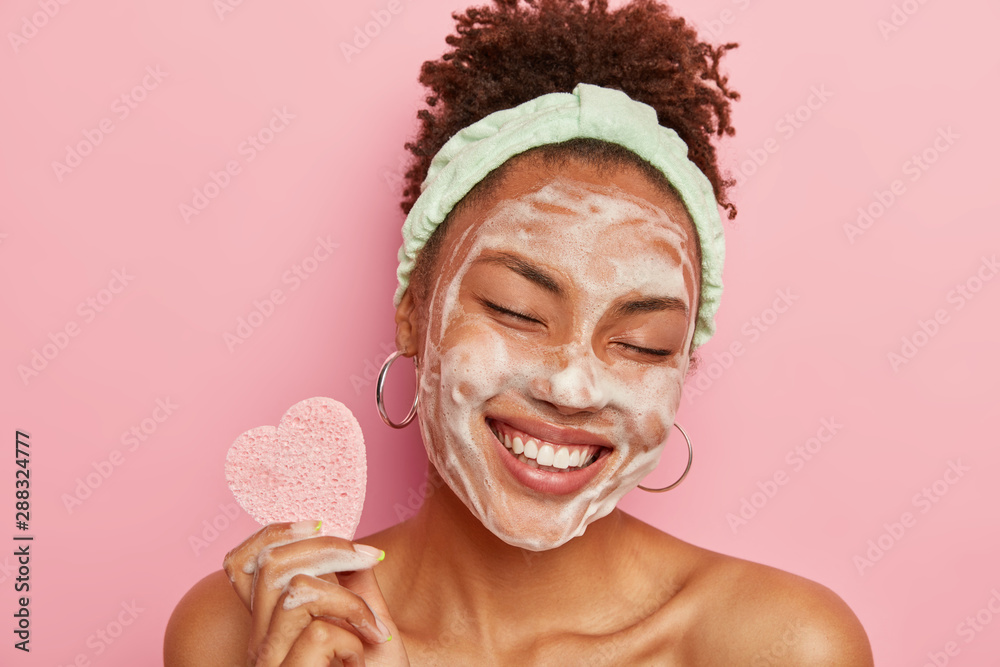 Beautiful optimistic Afro American woman cleanses face with foam, refreshes skin, has well cared complexion, holds heart shaped sponge for beauty procedures, stands bare shoulders with closed eyes - obrazy, fototapety, plakaty 