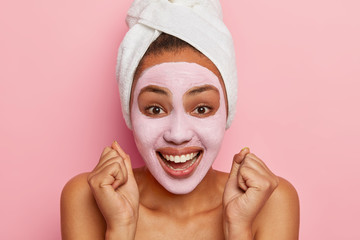 Wall Mural - Close up portrait of happy African American woman clenches fists, applies rosy clay mask, gets spa therapy, wears wrapped towel on wet hair, isolated over pink background, takes care of complexion