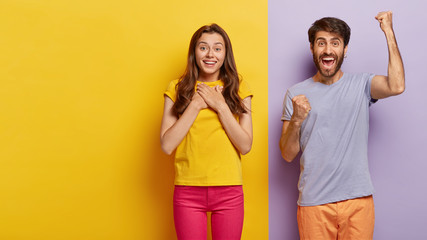 Wall Mural - Touched pleased young European woman keeps palms on chest, gets pleasure from hearing heart warming words, overjoyed man shouts yes and clenches fists, stand together against two colored background