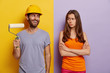 © wayhome.studio  - Happy hard working male in hardhat, holds paint roller, displeased wife stands crossed hands and looks at him, do house repairing at home, dressed in casual wear, isolated over two colored background
