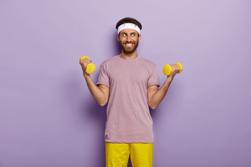 Indoor shot of happy man with bristle, raises two arms with weights, dressed in casual outfit, trains on biceps, poses against purple background. Motivated sportsman has workout in gym, healthy life