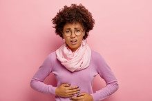 Young Afro American Woman Suffers From Painful Cramps In Belly, Has Stomachache, Frowns Face From Pain, Wears Round Spectacles, Purple Jumper With Scarf And Earrings, Being Hungry, Feels Ache