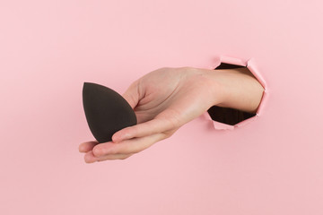 Wall Mural - Girl hand holds a beauty blender for makeup from a hole in a pink background, copy space.