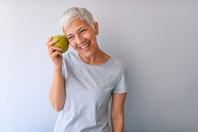 Portrait Of A Beautiful Elderly Woman Holding An Apple, Smiling, Isolated On Gray Background. Happy Woman Holding Granny Smith Apple. An Aple A Day Keeps Doctor Away